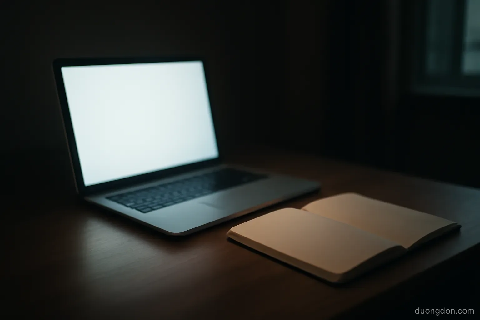 a computer screen glow reflecting on a desk in a dim room with a notebook open beside it, screen facing away, no readable content